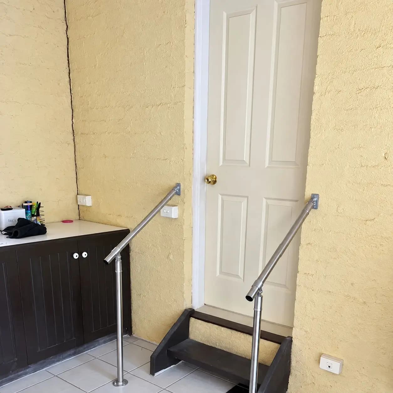 After: two stainless steel handrails, one on each side of the two steps up to an internal doorway, each fixed to the wall at the top and standing on a floor post at the bottom