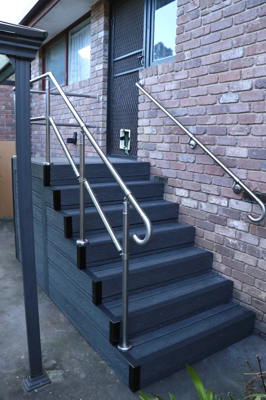 Stainless steel handrails on both sides of a set of dark composite entry steps against a brick wall